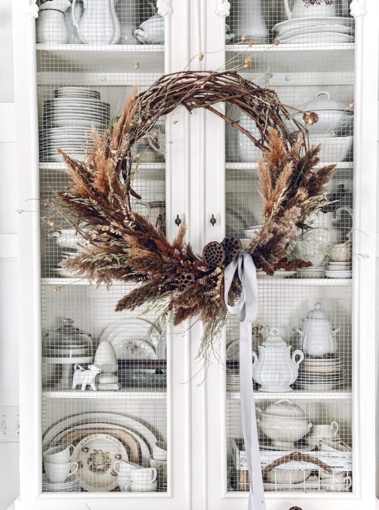 A brown and tan wreath with a white bow hanging on a china cabinet 
